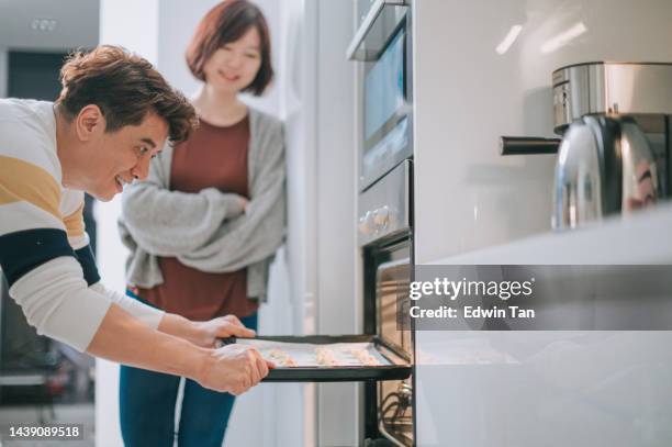 asian chinese couple making cookies inserting tray of cookies into oven at home in kitchen during weekend - inserting imagens e fotografias de stock