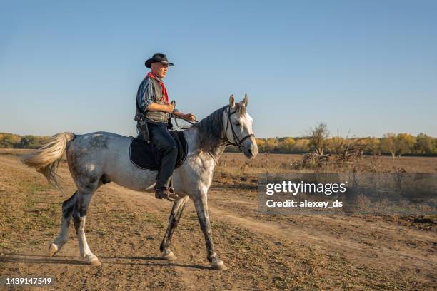 man wearing cowboy outfit goes on horseback on white horse in country road - western shirt stockfoto's en -beelden