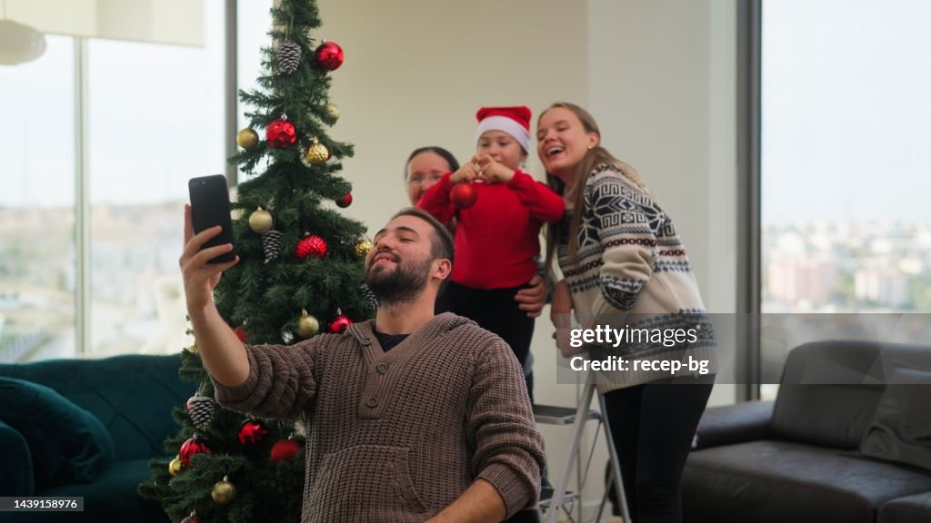 Young father taking photos and videos with smart phone of his family while decorating Christmas tree in living room at home