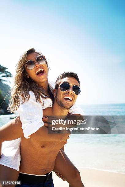 cheerful couple, man piggyback a beautiful woman on the beach. - couple on beach sunglasses stock pictures, royalty-free photos & images