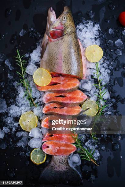 raw fresh salmon fish on ice cubes decorated with lemon slices, herbs and vegetables on black wooden background - frozen fish fillets stockfoto's en -beelden