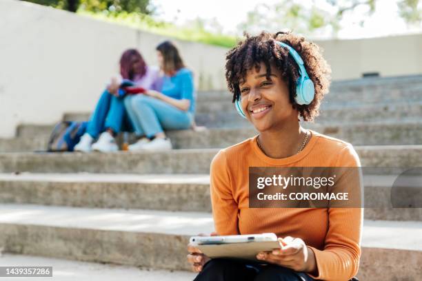 joven mujer latina escuchando música con unos auriculares azules sentada en unas escaleras sosteniendo la tableta digital - mujer sentada stock-fotos und bilder