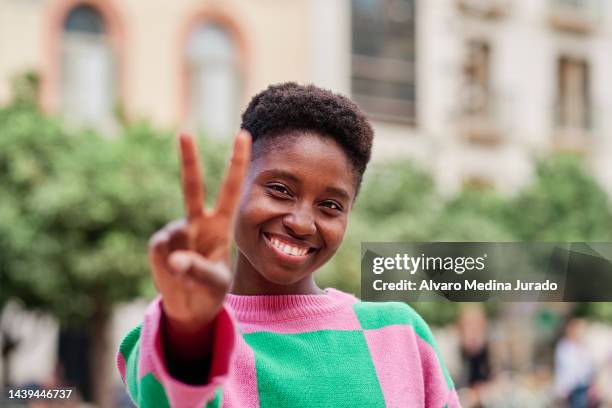 front view of a cheerful young african american woman making the v gesture, showing two fingers of her hand to the camera. - friedenszeichen handzeichen stock-fotos und bilder
