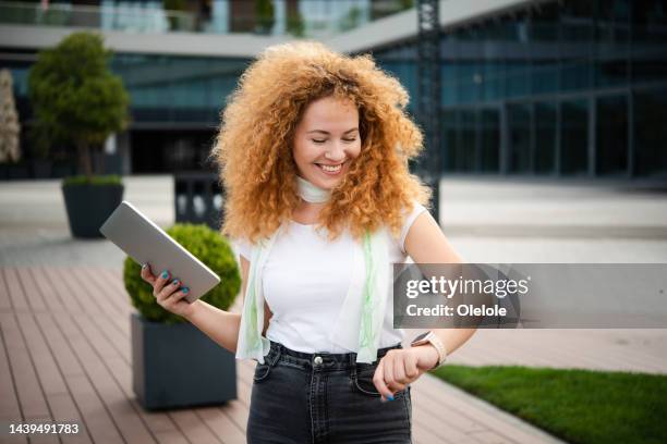 portrait of a beautiful redhead young woman checking the time and using tablet while walking - smart watch stock pictures, royalty-free photos & images