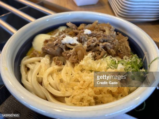 Bowl of Marugame's Niku Udon soup, from above, with tenkasu, sliced beef, udon noodles, and scallions presented in a blue and white pottery bowl, San...