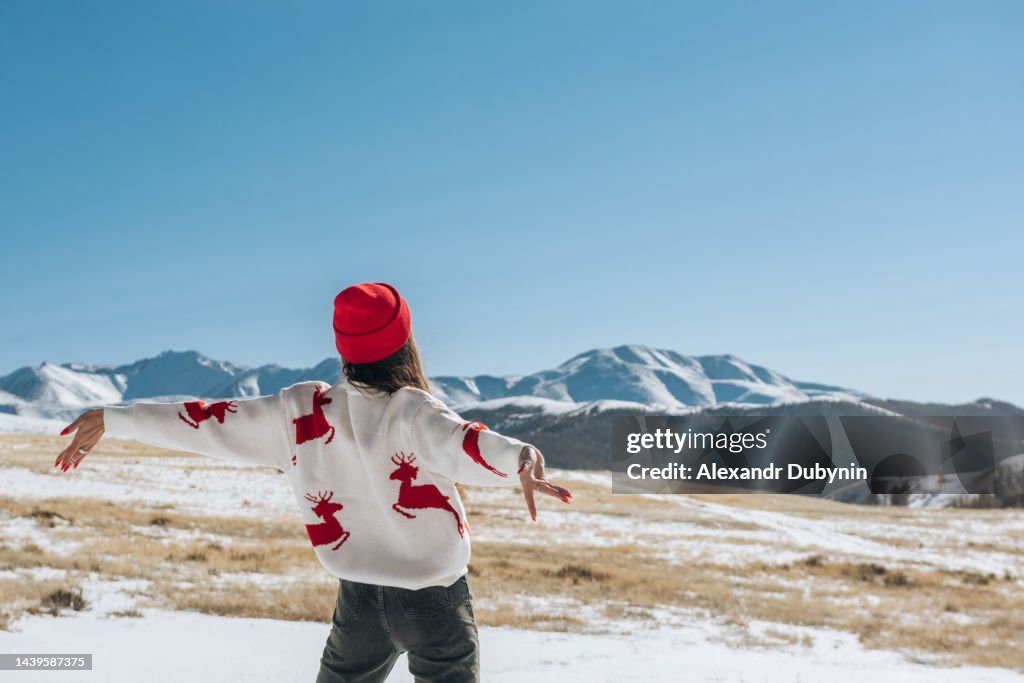 Happy young woman with raised hands on the background of blue sky and winter mountains in winter looking at the landscape. Christmas travel concept