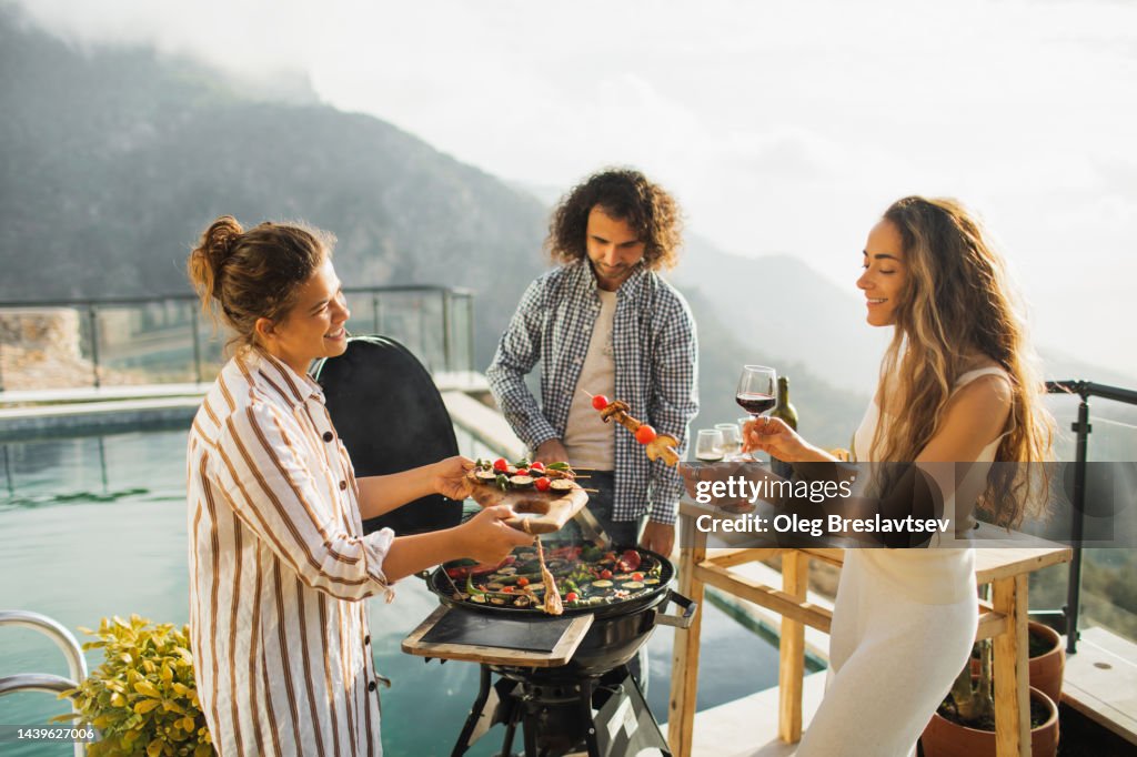 Friends cooking on grill and drinking wine outdoors on poolside with mountain view