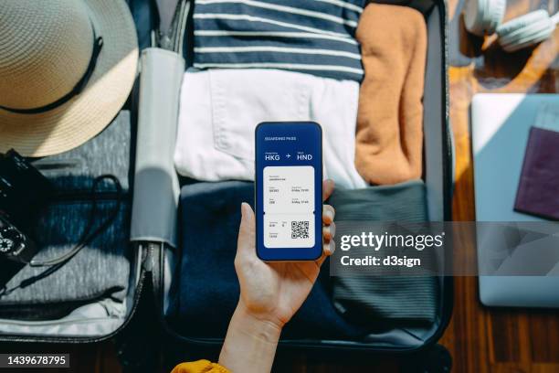 flat lay of woman's hand holding smartphone showing electronic flight ticket against packed suitcase with clothings, sunhat, camera, laptop and passport on wooden floor against sunlight. traveller's essentials. travel and vacation concept. business travel - travel essentials stock pictures, royalty-free photos & images