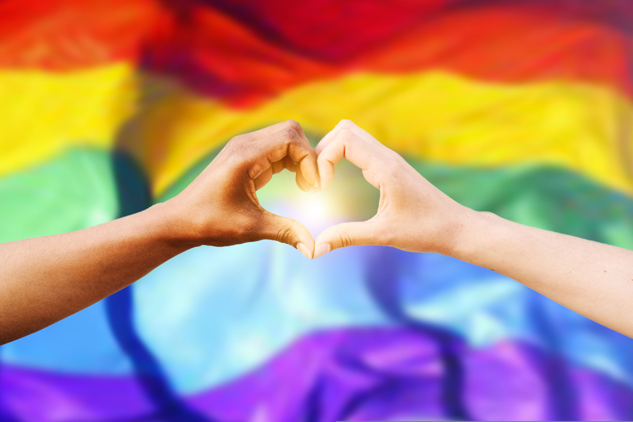 Two multiracial hands join together to form a shape heart with a LGBT flag background. Concept of homosexual gay pride day and love between different races, peace, humans against discrimination Two multiracial hands join together to form a shape heart with a LGBT flag background. Concept of homosexual gay pride day and love between different races, peace, humans against discrimination