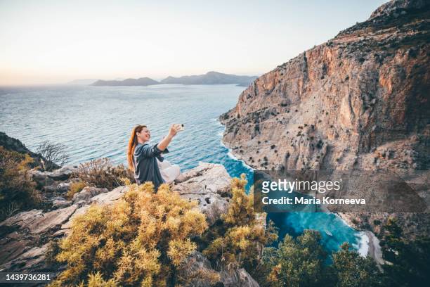 young woman sitting on rocks and making selfie at beautiful sea view. - tourist selfie stock pictures, royalty-free photos & images