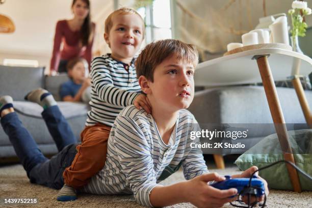 a toddler sits on the back of his brother who is lying on the carpet in the living room and plays video games - game three stockfoto's en -beelden