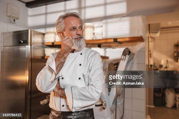panadero maduro con los brazos ligeramente cruzados de pie en la cocina de la panadería - uniforme de chef fotografías e imágenes de stock