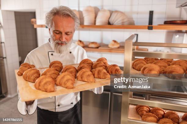 un boulanger caucasien mature regardant et tenant un plateau rempli de croissants fraîchement cuits - baker smelling bread photos et images de collection