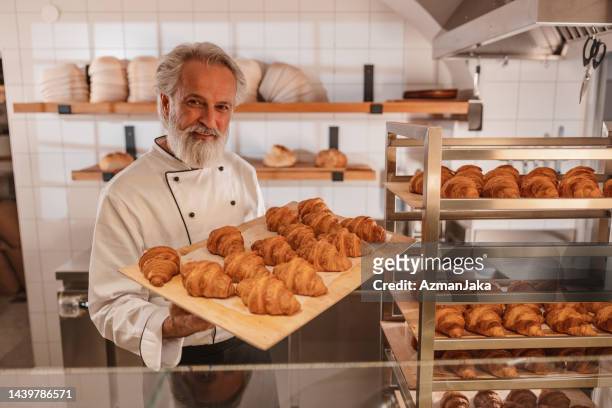happy mature baker regardant et tenant un plateau plein de croissants fraîchement cuits - baker smelling bread photos et images de collection