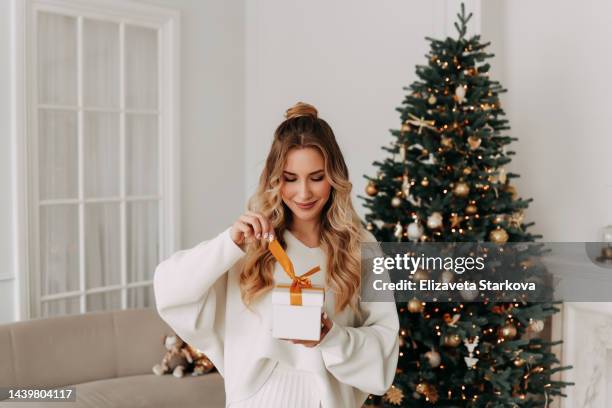 the concept of christmas. portrait of a smiling young woman with curly hair in a knitted sweater holding and opening a christmas gift on the background of a christmas tree in a bright interior on a holiday at home - when travel was a thing of style stockfoto's en -beelden