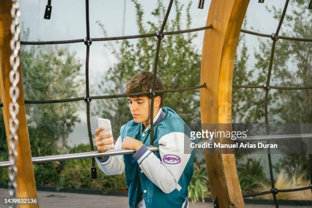 portrait of a handsome young man in a blue jacket looking at his phone leaning against a metal structure in a playground. - felicidad duran fotografías e imágenes de stock