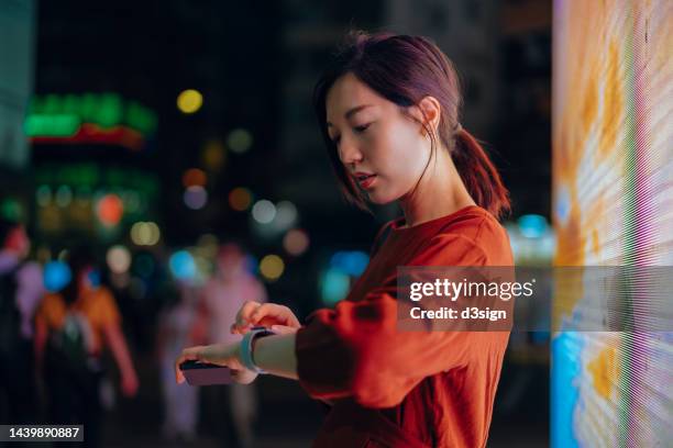 young asian woman with smartphone using smartwatch in city street at night. standing in front of colourful and illuminated led digital display. city scene in bokeh lights in background. lifestyle and technology. interconnections of the internet - asian and indian ethnicities smartwatch phone stock pictures, royalty-free photos & images