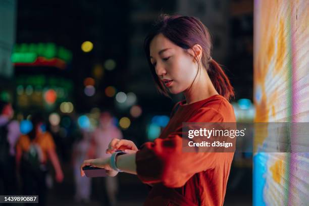 young asian woman with smartphone using smartwatch in city street at night. standing in front of colourful and illuminated led digital display. city scene in bokeh lights in background. lifestyle and technology. interconnections of the internet - digital payment imagens e fotografias de stock