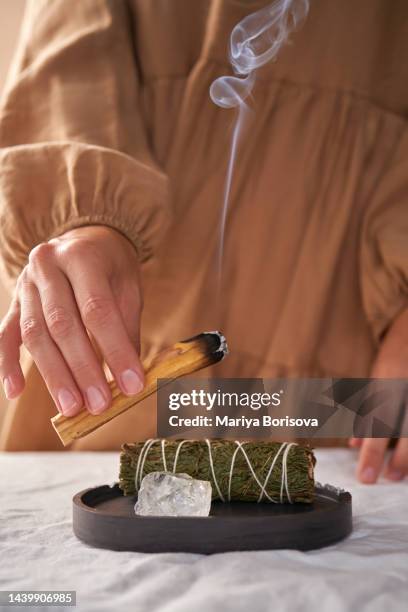 the hands of a girl in a beige dress hold a smoking palo santo stick against the background of ritual things: a rock crystal stone and a twist for fumigation. - fumigation photos et images de collection