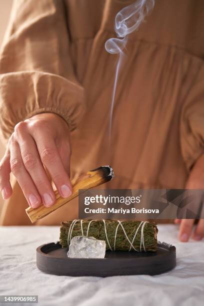 the hands of a girl in a beige dress hold a smoking palo santo stick against the background of ritual things: a rock crystal stone and a twist for fumigation. - smudging ceremony stock pictures, royalty-free photos & images