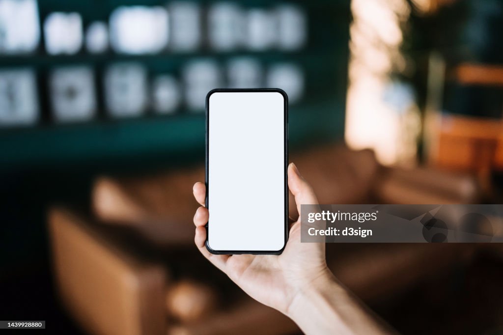 Close up, cropped shot of a woman's hand holding up a smartphone with blank white screen in a cozy and stylish living room interior. Lifestyle and technology. Smartphone with blank screen for design mockup
