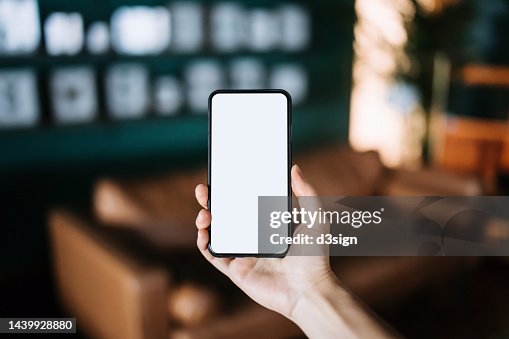 Close up, cropped shot of a woman's hand holding up a smartphone with blank white screen in a cozy and stylish living room interior. Lifestyle and technology. Smartphone with blank screen for design mockup Close up, cropped shot of a woman's hand holding up a smartphone with blank white screen in a cozy and stylish living room interior. Lifestyle and technology. Smartphone with blank screen for design mockup