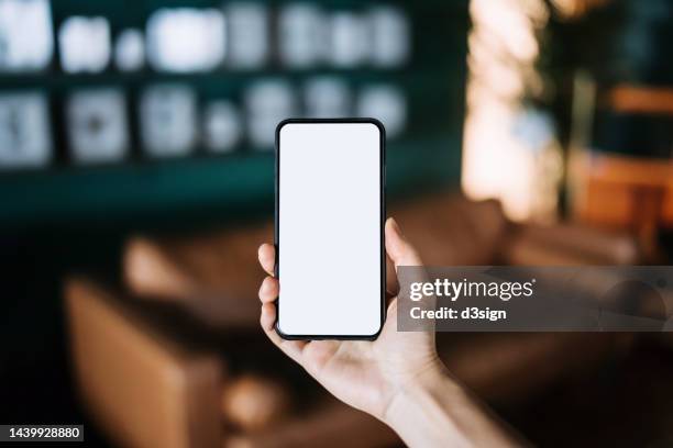 close up, cropped shot of a woman's hand holding up a smartphone with blank white screen in a cozy and stylish living room interior. lifestyle and technology. smartphone with blank screen for design mockup - phone blank screen stock pictures, royalty-free photos & images