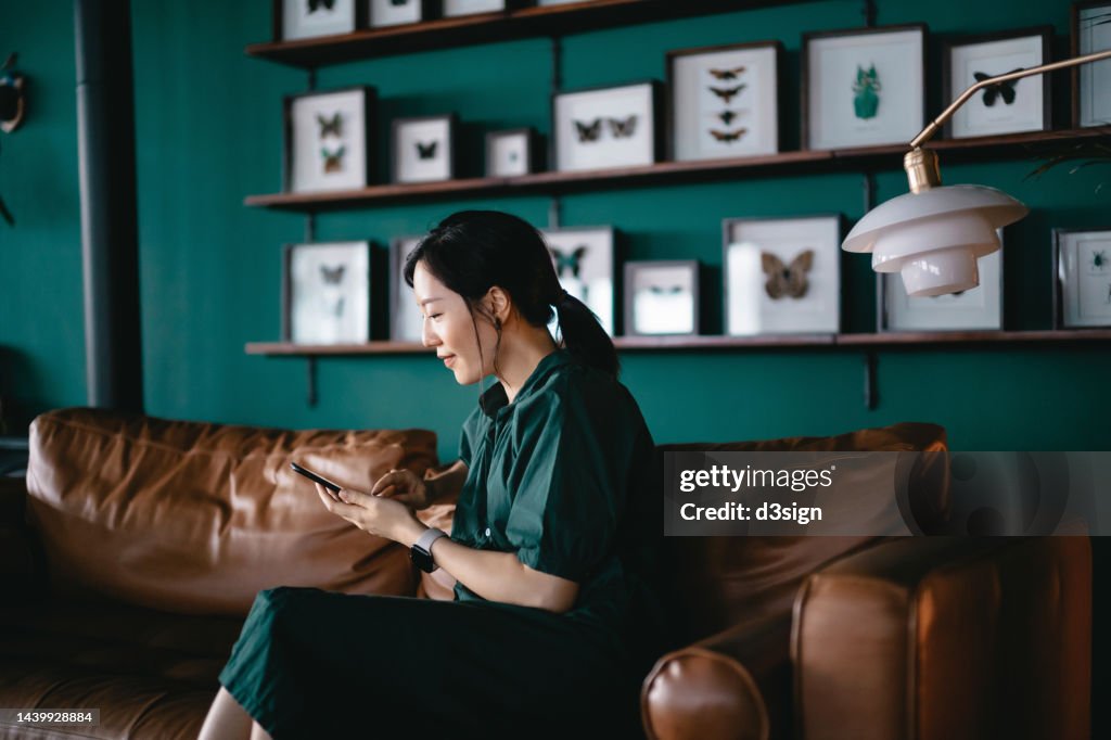 Beautiful smiling young Asian woman managing personal banking and finance with online banking mobile app on smartphone, sitting on sofa in the living room at cozy home. Wealth management. Digital banking habits. Smart banking with technology