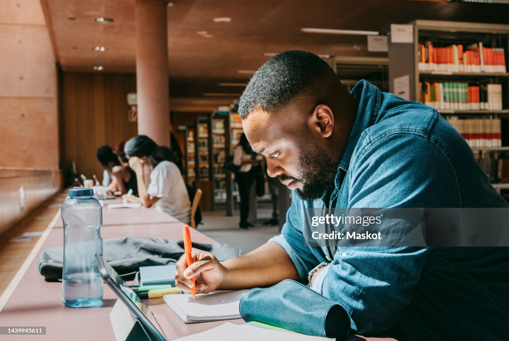 Student writing in notebook while sitting with tablet PC in college library
