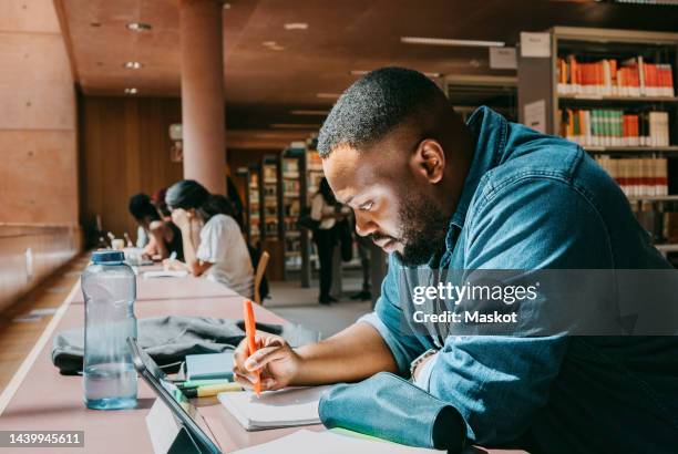 student writing in notebook while sitting with tablet pc in college library - student stockfoto's en -beelden