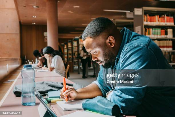 student writing in notebook while sitting with tablet pc in college library - college education stock pictures, royalty-free photos & images