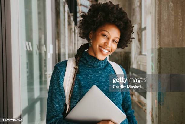 smiling female student with laptop and curly hair standing in corridor - toothy smile stock pictures, royalty-free photos & images