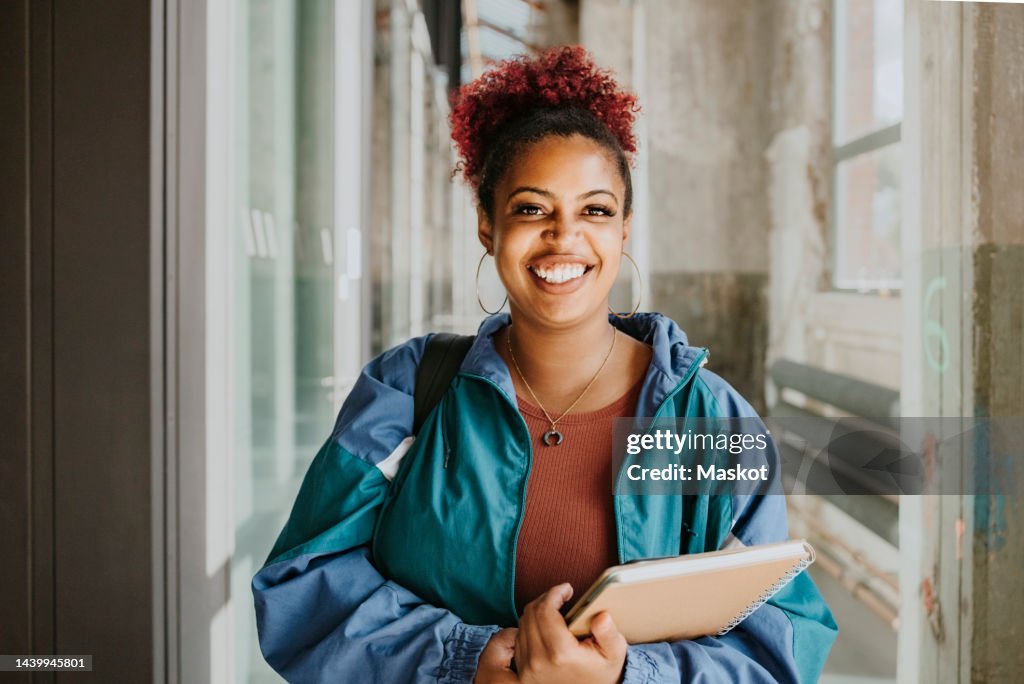 Portrait of happy female student wearing jacket holding books in college corridor