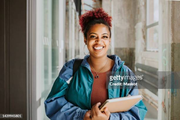 portrait of happy female student wearing jacket holding books in college corridor - edifício público - fotografias e filmes do acervo