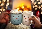 Mom and child are relaxing together on a cozy winter evening by the fireplace, close-up of two hands with snowman face cup of hot cocoa with marshmallows. Christmas holidays, happy moments at home.