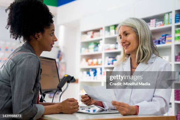 mature female pharmacist taking an african-american woman's prescription order while standing behind pharmacy's counter - black pharmacist stock pictures, royalty-free photos & images