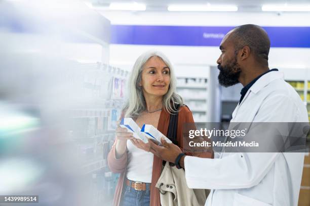 mid-shot of male african-american pharmacist assisting a middle-aged female customer while standing in pharmacy's aisle - cosmetics counter stock pictures, royalty-free photos & images