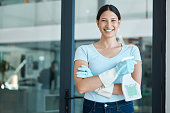 Cleaning service, portrait and cleaner in an office with spray bottle of disinfectant, bleach or detergent. Happy, smile and young female worker with gloves and soap liquid done washing glass windows