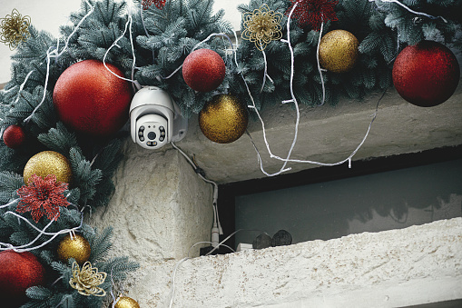 Stylish safety camera and christmas baubles on fir branches on building exterior. Modern christmas decor and security equipment in city street. Winter holidays in Europe.