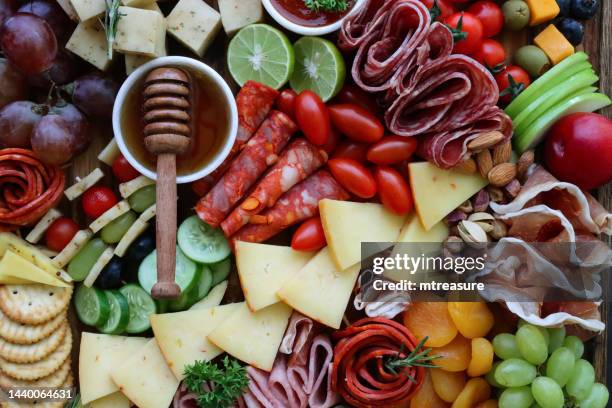 full frame image of charcuterie board, apricots, crackers, ramekin of honey, salami roses, red and white grapes, cherry vine tomatoes, apple slices, cheddar and herb cheese, elevated view - cutting board stock pictures, royalty-free photos & images