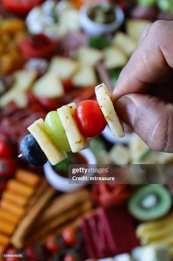 Close-up image of unrecognisable person holding cheese and fruit cocktail stick kebab over charcuterie board, crackers, ramekin of chutney, salami roses, red grapes, cherry vine tomatoes, kiwi half, cheddar and herb cheese cubes, elevated view