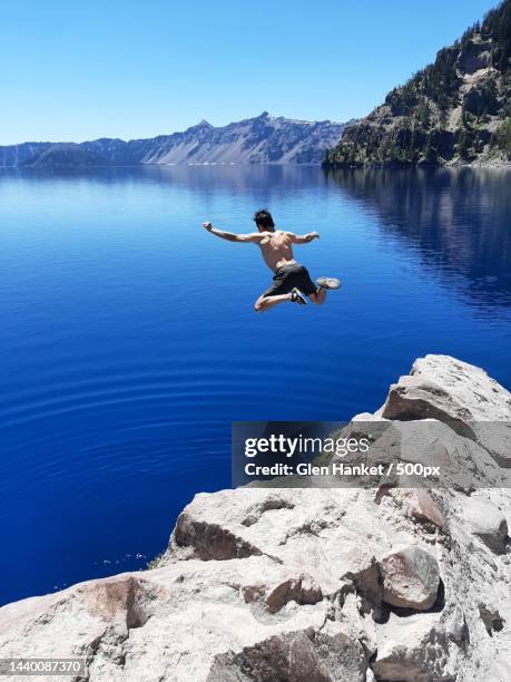 man jumping in lake against clear blue sky - salto desde acantilado fotografías e imágenes de stock