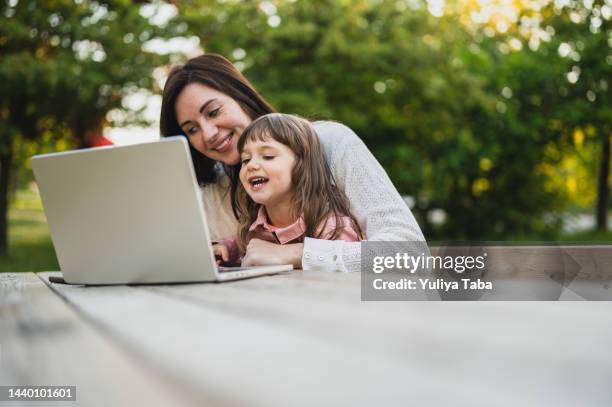 mutter, die am laptop arbeitet, während sie sich mit ihrer tochter in einem öffentlichen park ausruht und entspannt. - familie laptop stock-fotos und bilder