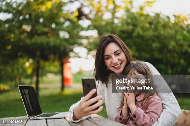 jovem mãe e sua filha têm chamada de vídeo enquanto descansam em um parque. mãe usando laptop. - 2 3 anos - fotografias e filmes do acervo