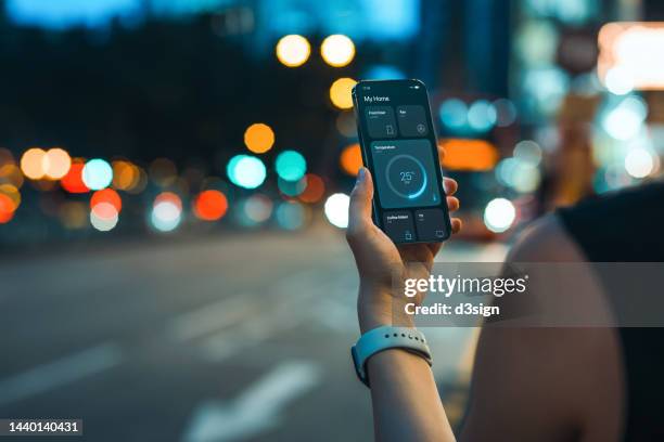 over the shoulder view of young woman turning on the air-conditioner in her smart home with home automation mobile app on smartphone on her way home. home automation. smart living. lifestyle and technology. smart home technology concept - application modernization stock pictures, royalty-free photos & images