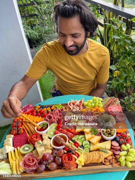 close-up image of man sat in front of charcuterie board on apartment balcony table, ramekins of honey and chutney, crackers, salami roses, breadsticks, kiwi, vines tomatoes, soft and hard cheeses, stuffed olives, blueberries, elevated view - leicester cheese stock pictures, royalty-free photos & images