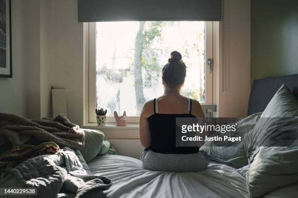 rear view of a teenage girl looking out of the window whilst sitting on her bed - desequilibrio fotografías e imágenes de stock