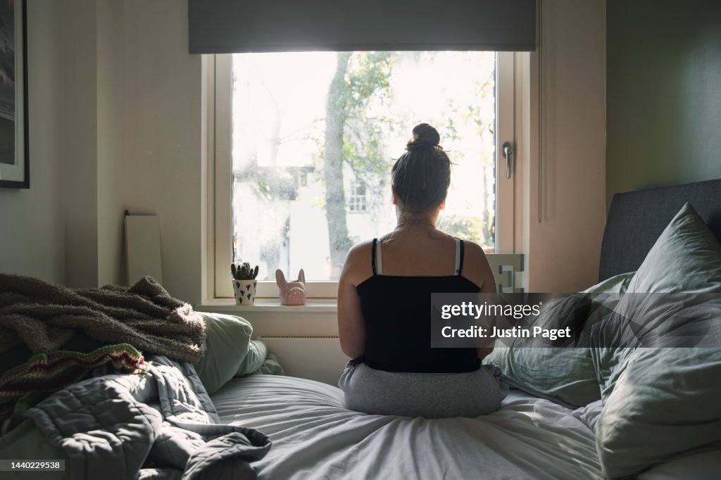 Rear view of a teenage girl looking out of the window whilst sitting on her bed