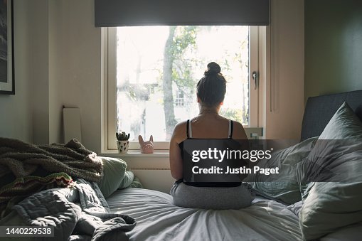 Rear view of a teenage girl looking out of the window whilst sitting on her bed Rear view of a teenage girl looking out of the window whilst sitting on her bed