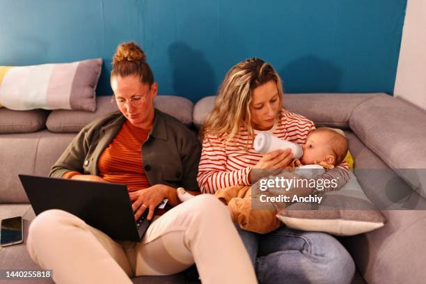 mujer alimentando con biberón a un bebé mientras su esposa trabaja en una computadora portátil - queer fotografías e imágenes de stock